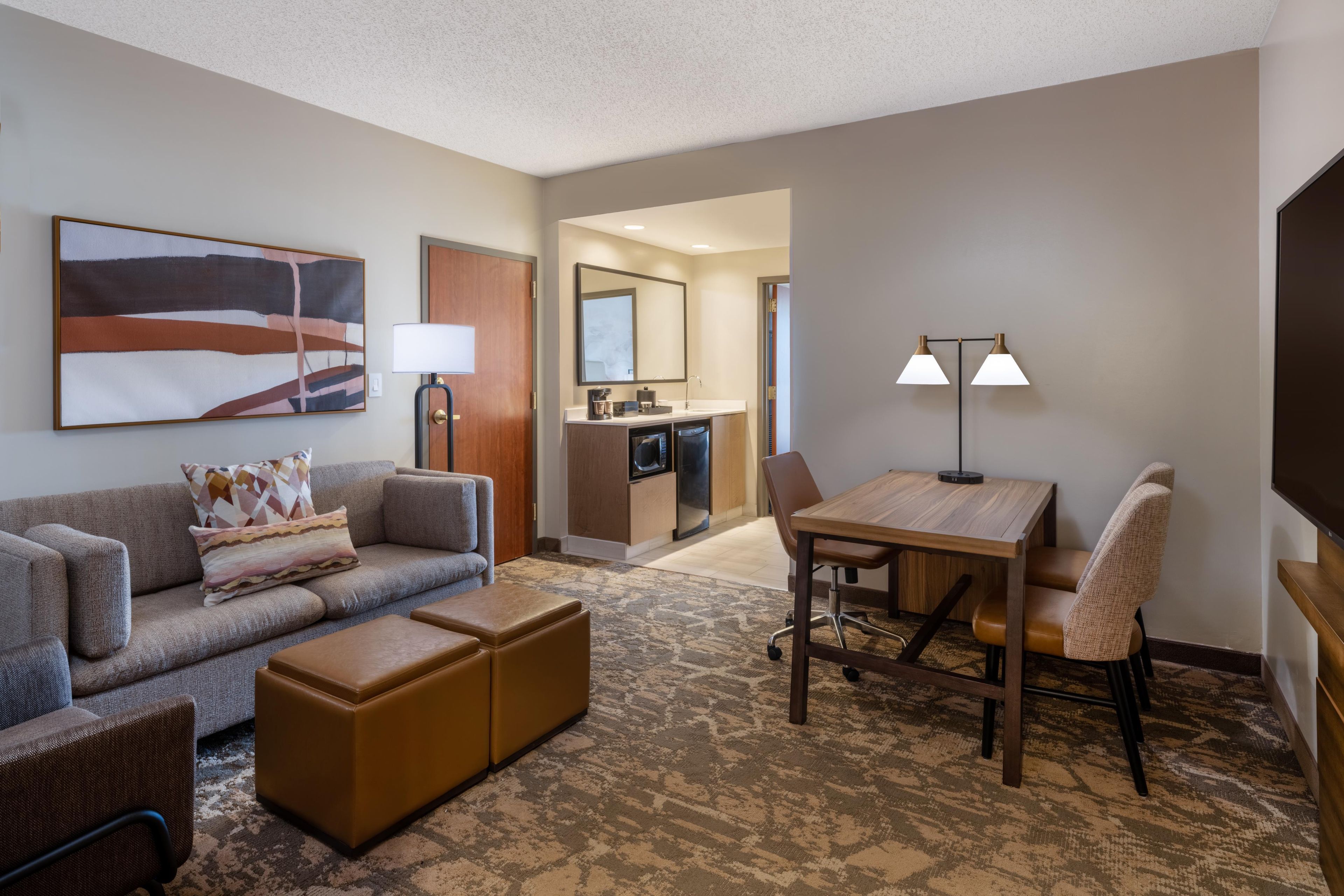 A hotel room with a sofa, ottoman, a dining table with chairs, a TV, a painting, and a kitchenette.