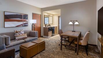 A hotel room with a sofa, ottoman, a dining table with chairs, a TV, a painting, and a kitchenette.