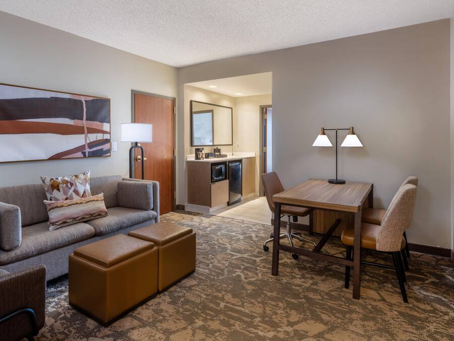 A hotel room with a sofa, ottoman, a dining table with chairs, a TV, a painting, and a kitchenette.