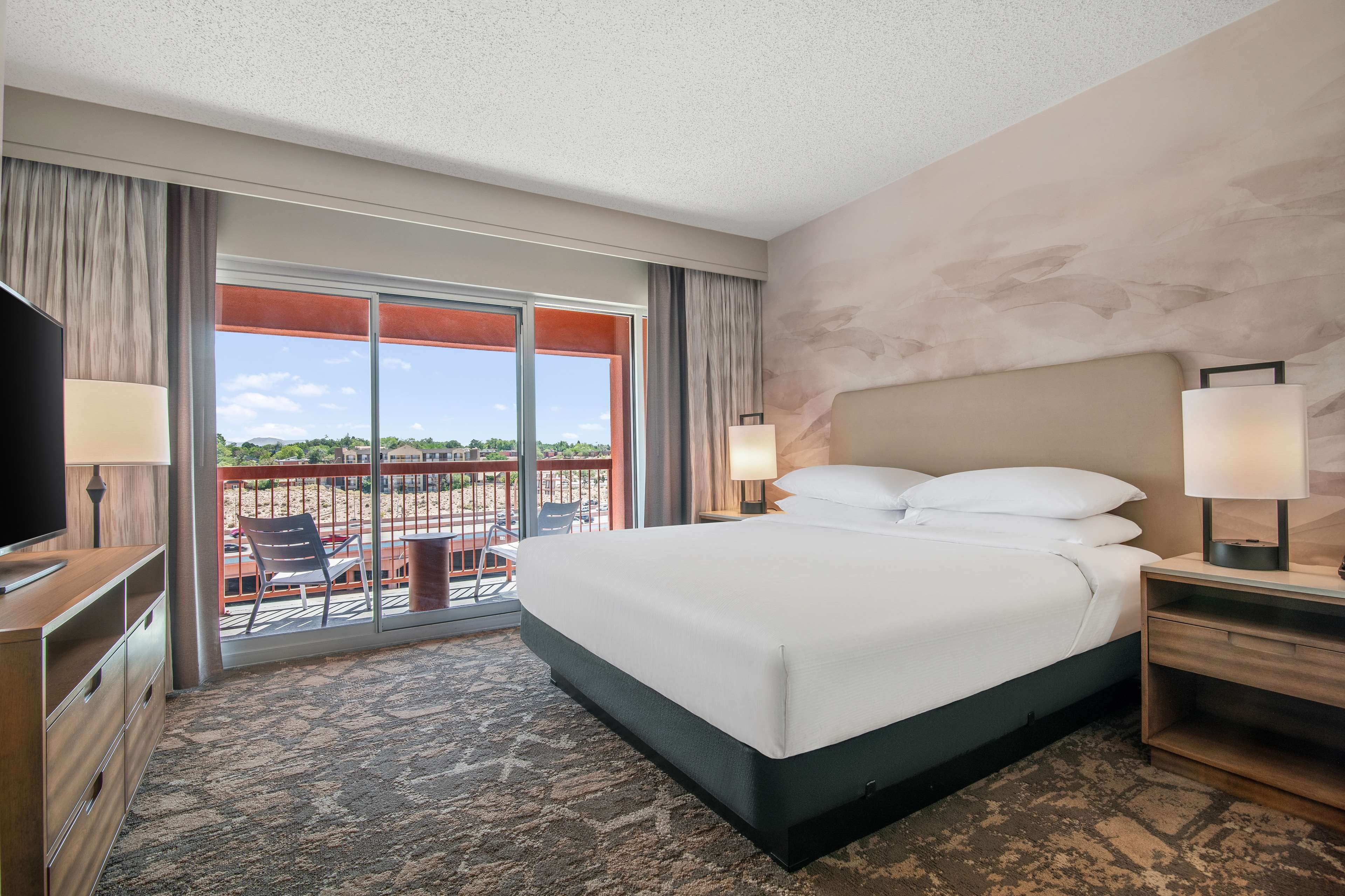 A hotel room with a large bed, two bedside tables with lamps, a TV, a balcony with a chair, and a view of the outdoors.