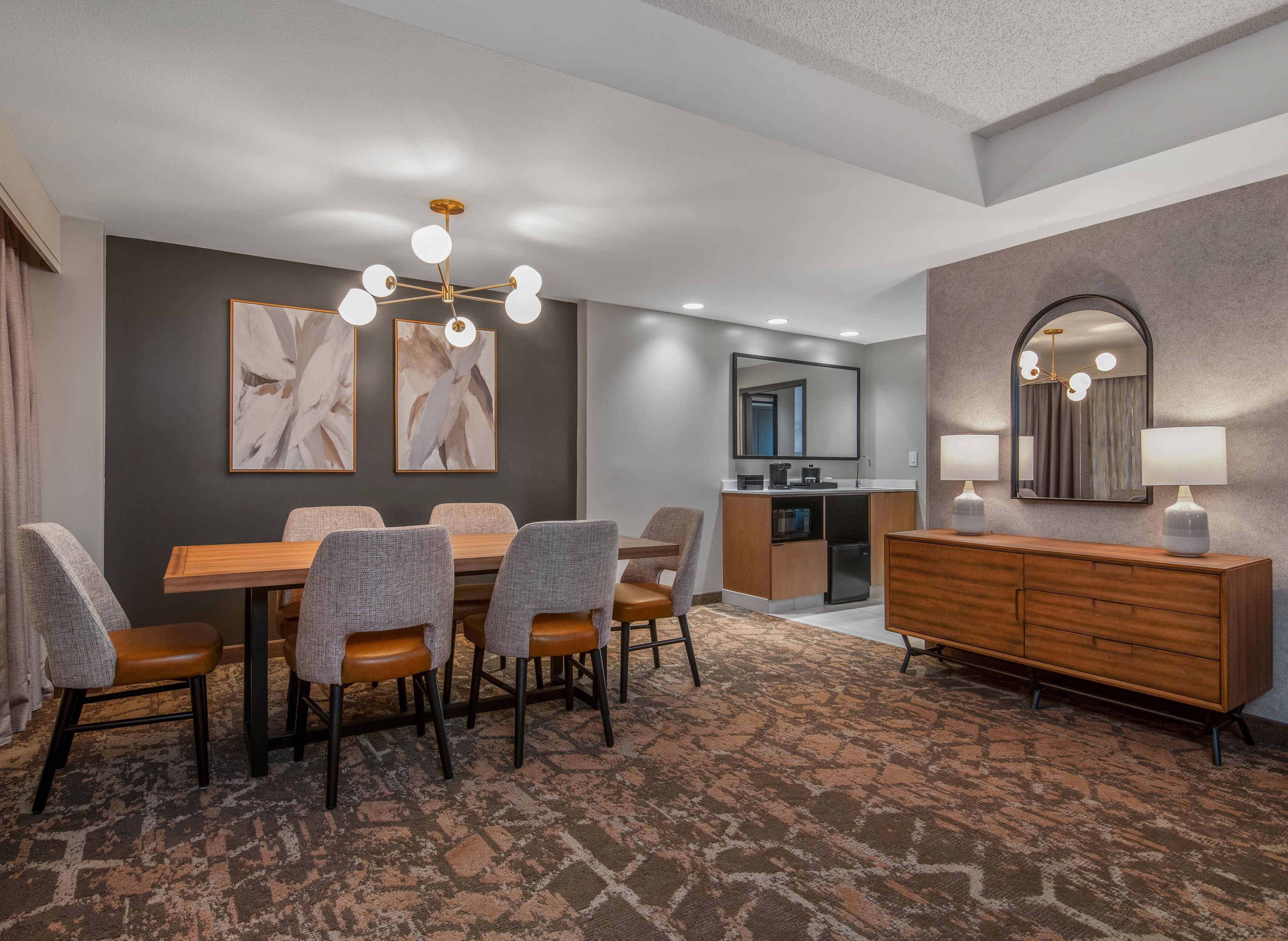 Habitación de hotel con comedor, kitchenette, espejo grande y alfombra estampada.