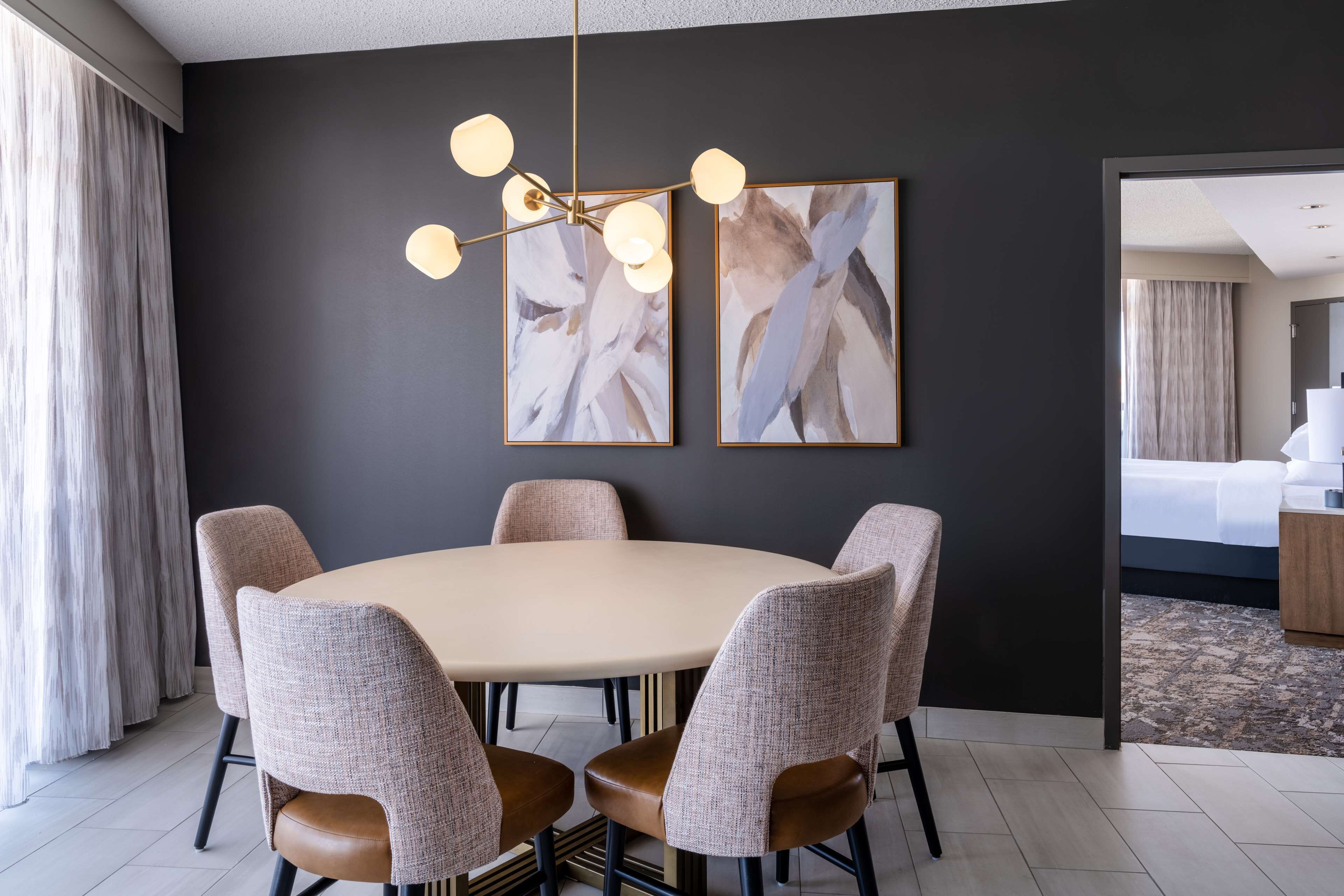 A modern hotel room with a round dining table, six upholstered chairs, a chandelier, abstract artwork on the wall, and a view into the bedroom.