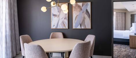 A modern hotel room with a round dining table, six upholstered chairs, a chandelier, abstract artwork on the wall, and a view into the bedroom.