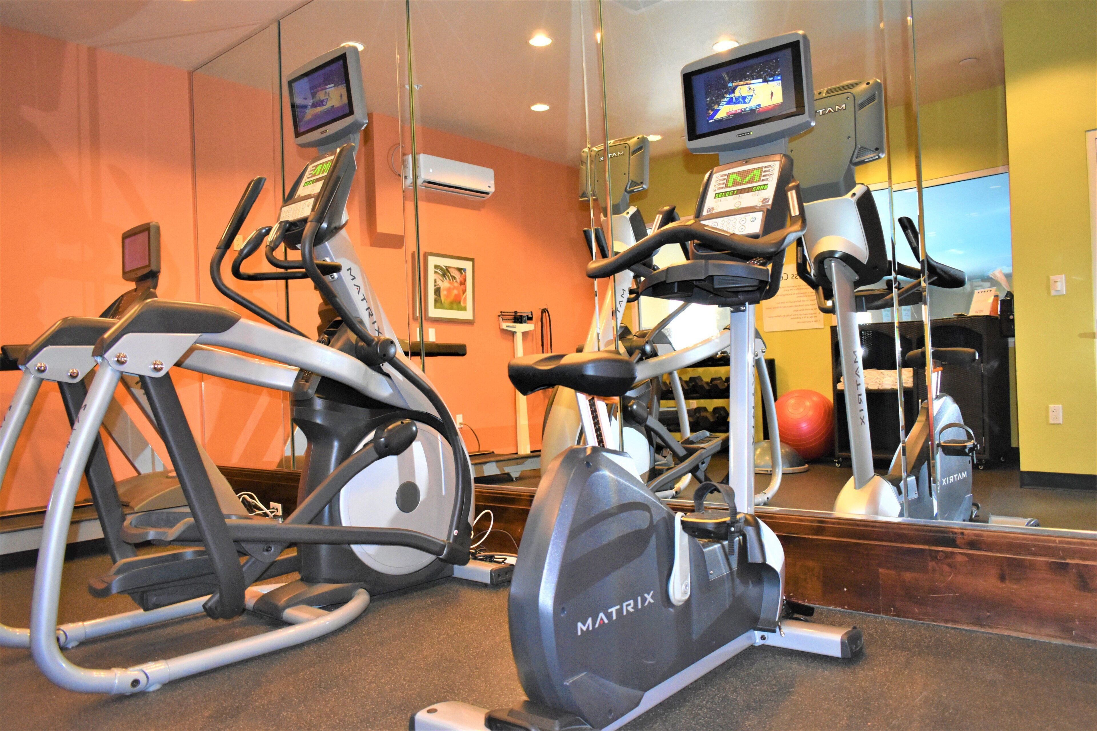 A gym with stationary bikes and a treadmill, a mirror reflecting the equipment, and a television screen mounted on the wall.