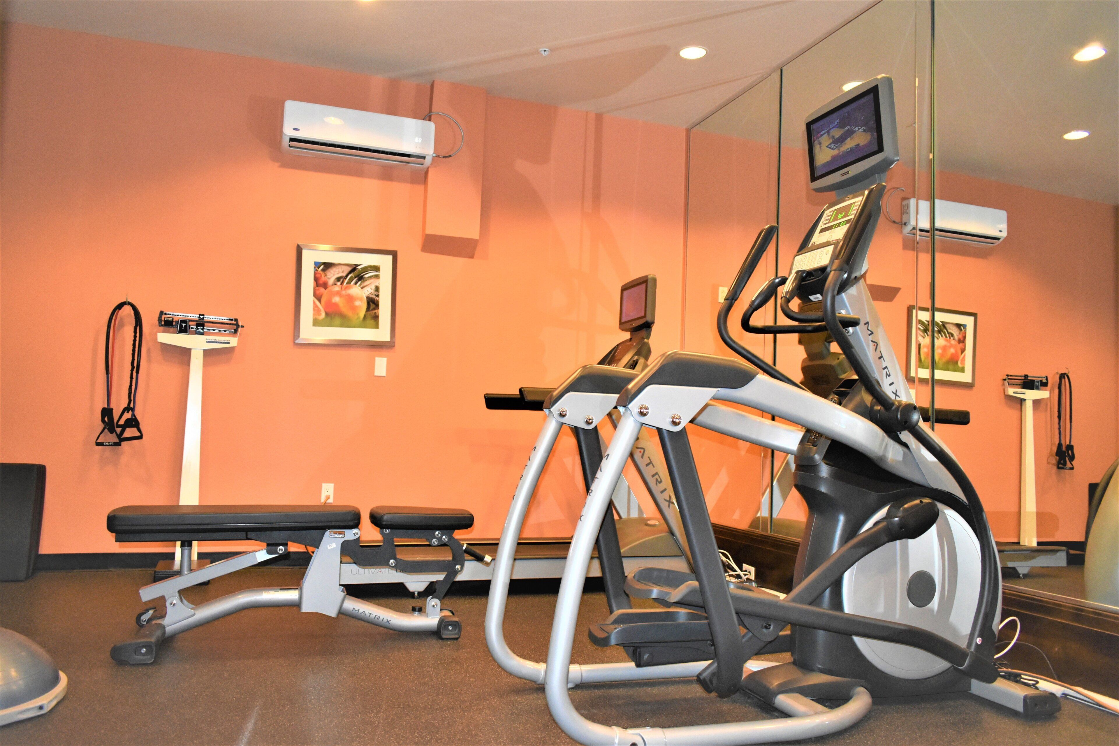 A gym with exercise equipment, including a treadmill and an elliptical machine, against an orange wall with framed pictures.