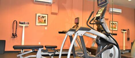 A gym with exercise equipment, including a treadmill and an elliptical machine, against an orange wall with framed pictures.
