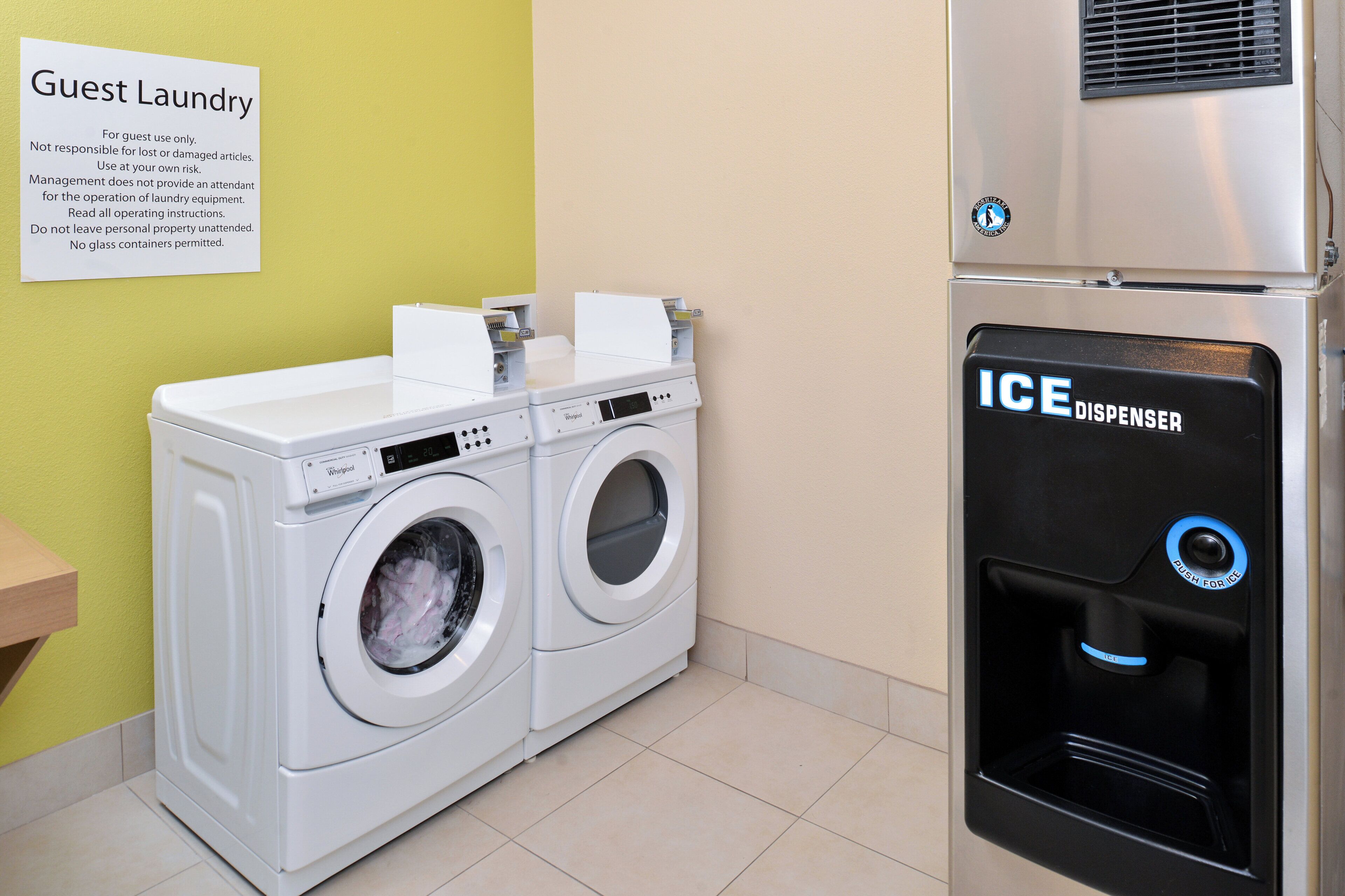 Uma área de lavanderia para hóspedes com duas máquinas de lavar Whirlpool de abertura frontal e um dispensador de gelo.