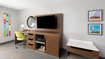 A modern hotel room with a wooden desk, a flat-screen TV, a yellow chair, a small coffee machine, and two framed pictures on the wall.