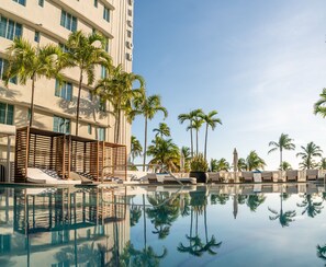 Un espace piscine d’hôtel où l’on peut admirer de beaux reflets, des palmiers et la façade moderne d’un immeuble.