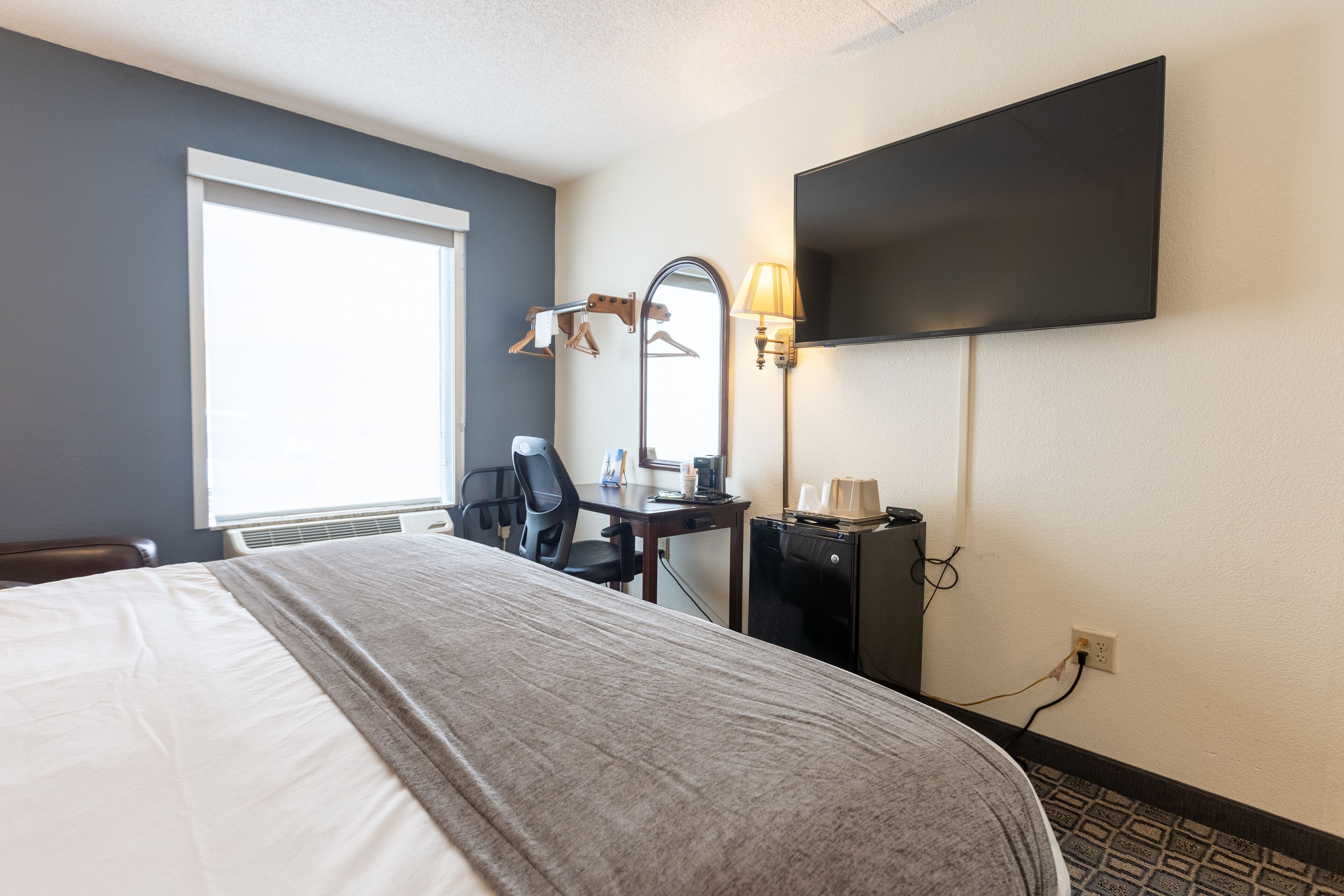 A hotel room with a large bed, a desk with a chair, a flat-screen TV mounted on the wall, and a window providing natural light.