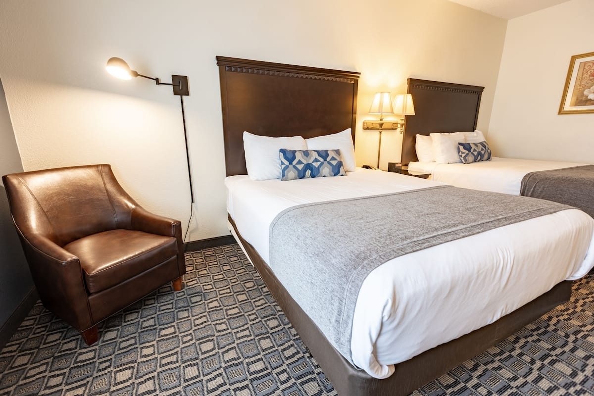 A hotel room with a brown leather armchair, two beds with white and gray bedding, a patterned floor, and a wall-mounted lamp.