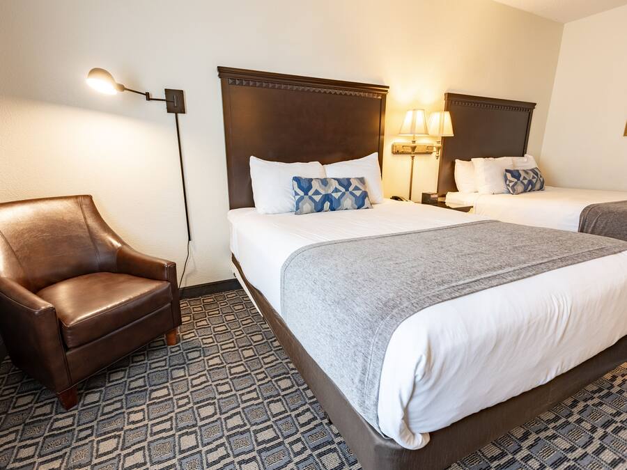 A hotel room with a brown leather armchair, two beds with white and gray bedding, a patterned floor, and a wall-mounted lamp.