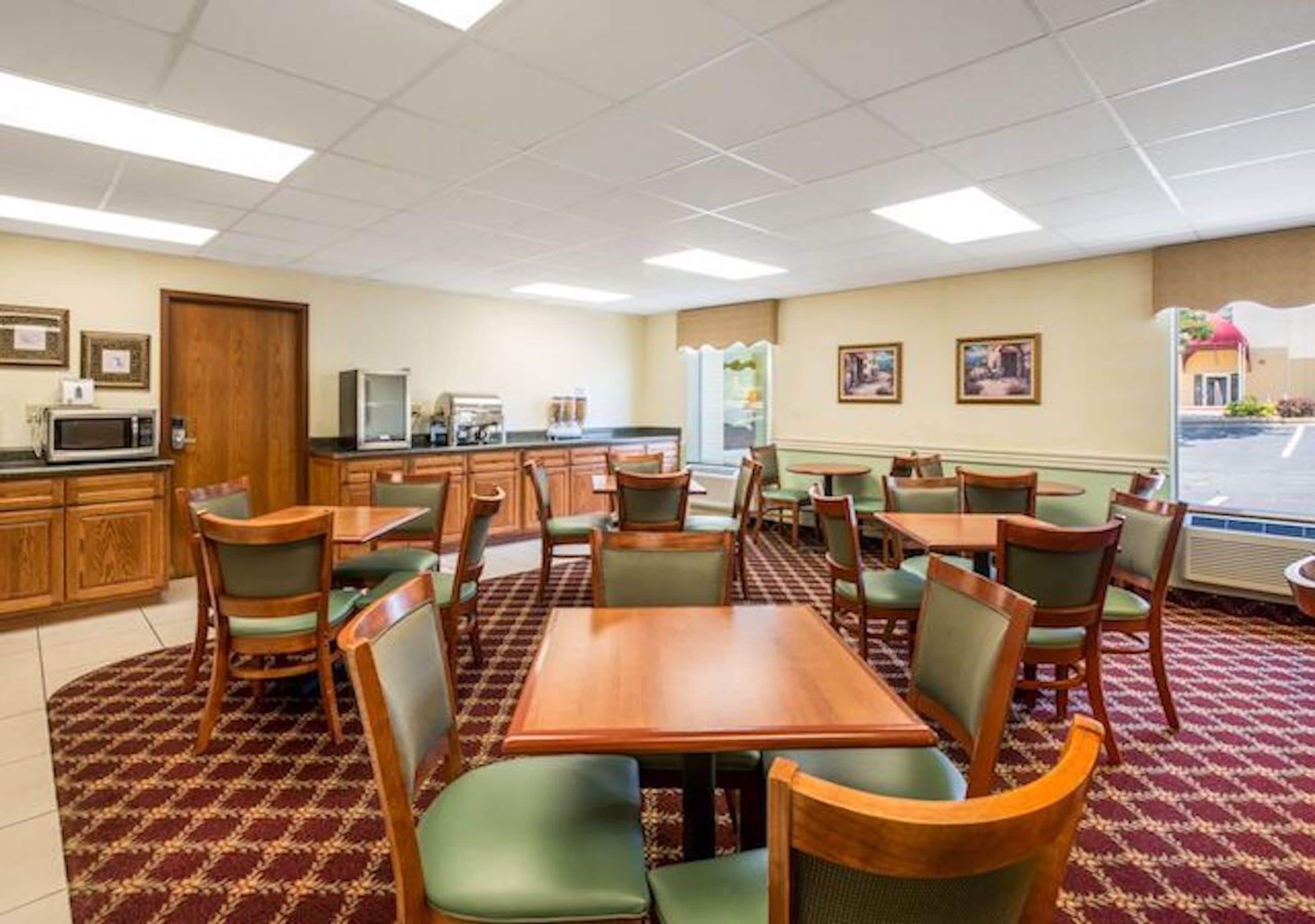 A hotel dining area with multiple tables and chairs, a kitchen area with appliances, and a view of the outside.