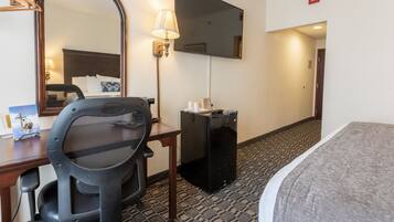 A hotel room with a bed, a desk with a chair, a mirror, a lamp, and a television.