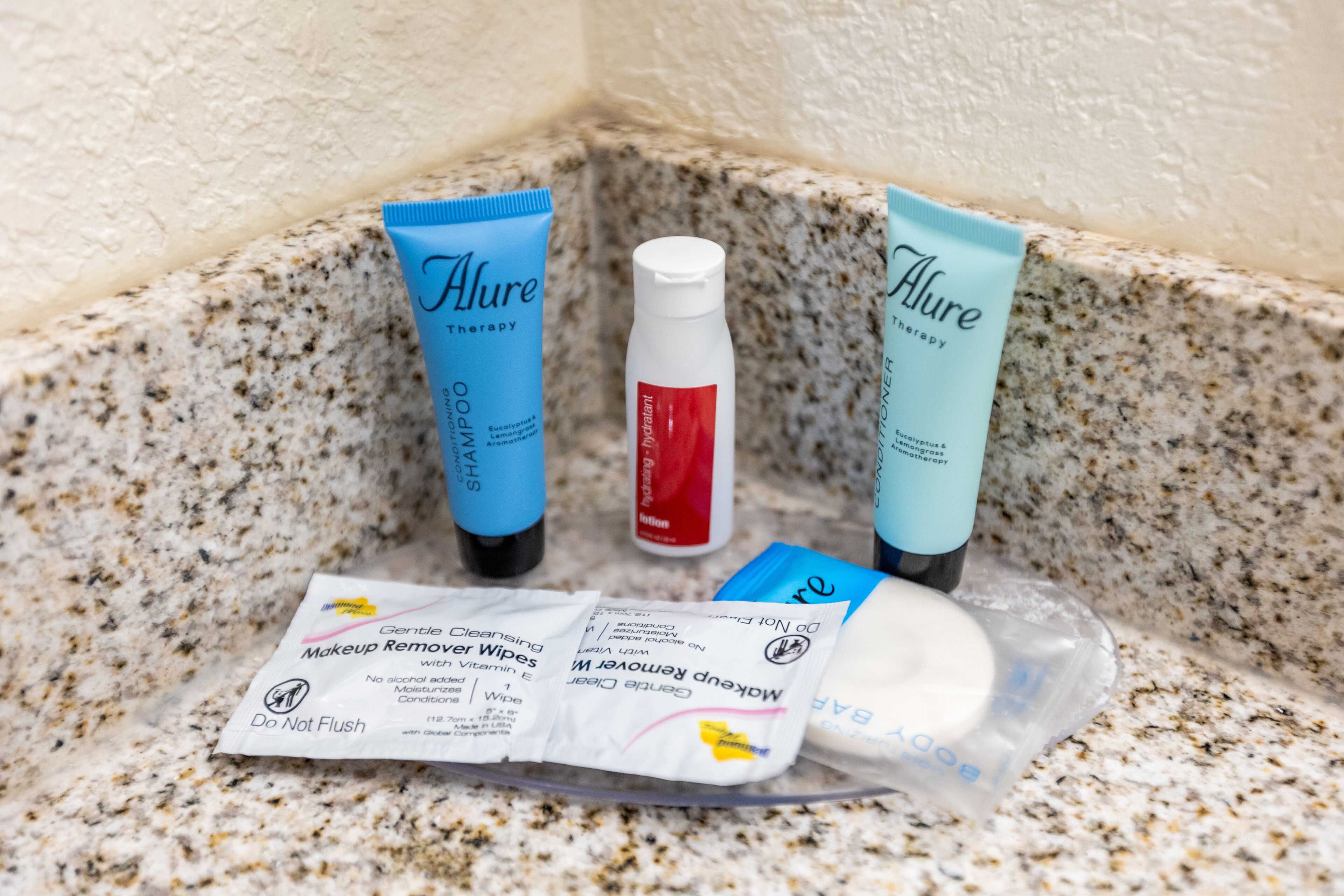 Un meuble-lavabo de salle de bain avec des produits de toilette, notamment deux tubes de shampooing « Alure Therapy », un flacon de lotion « Hydra-Lant » et un paquet de lingettes démaquillantes douces.