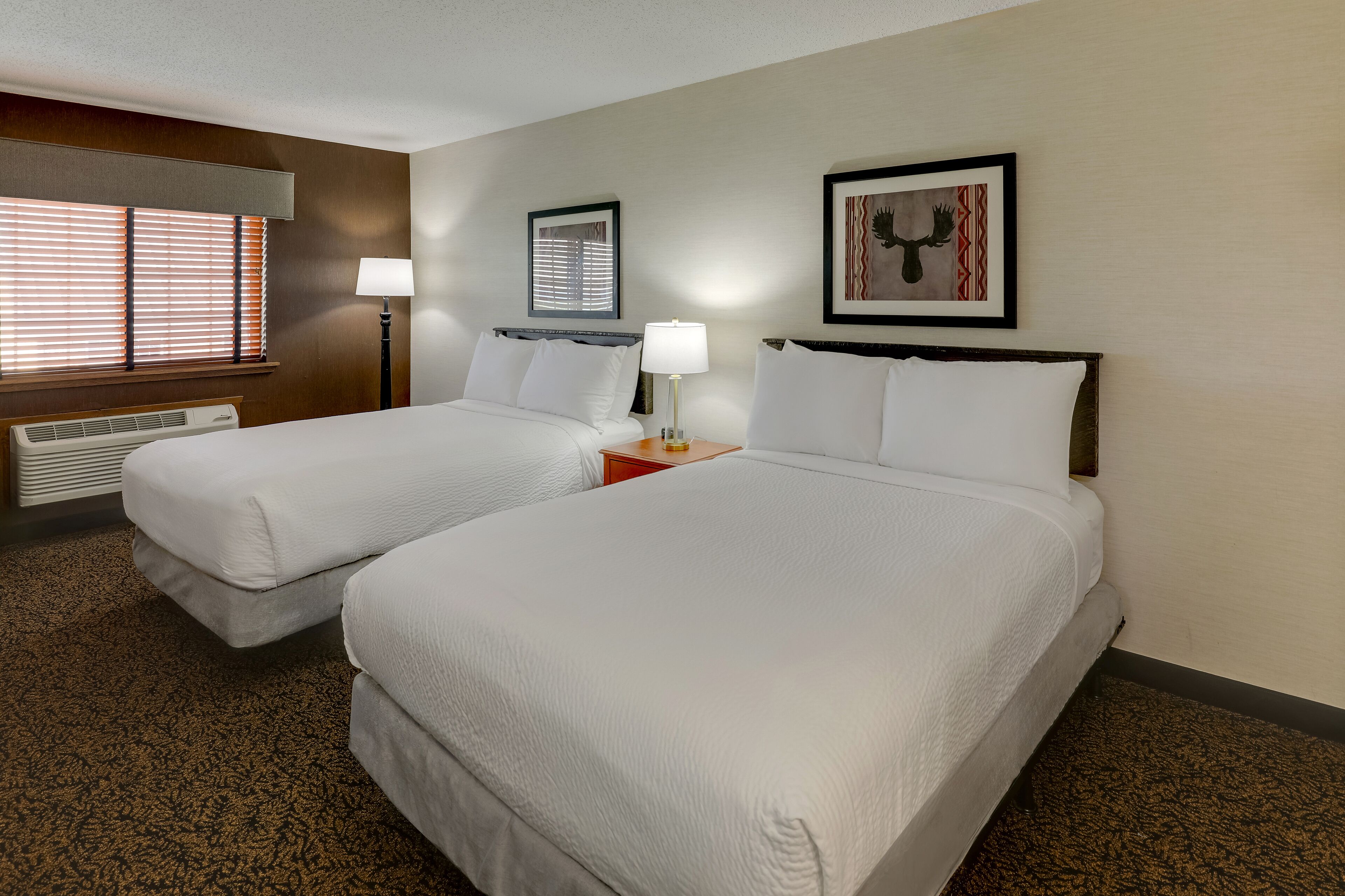 A hotel room with two beds, a window with blinds, a wall-mounted lamp, and a framed picture.