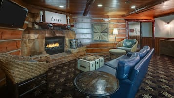 A cozy cabin interior with a stone fireplace, a television mounted on the wall, a wicker chair, a blue sofa, and a patterned rug.