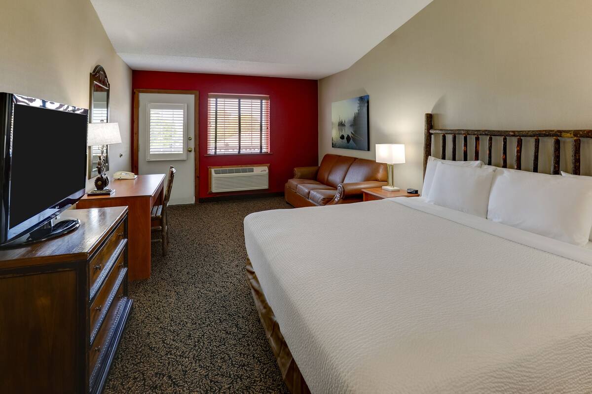Habitación de hotel con una cama grande, un escritorio con televisor, una silla, una pared de acento roja y una ventana con persianas.