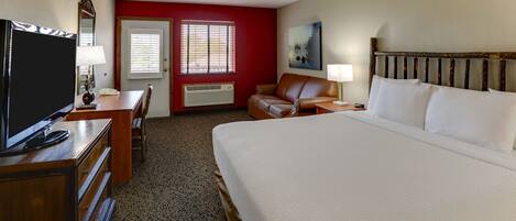 A hotel room with a large bed, a desk with a TV, a chair, a red accent wall, and a window with blinds.