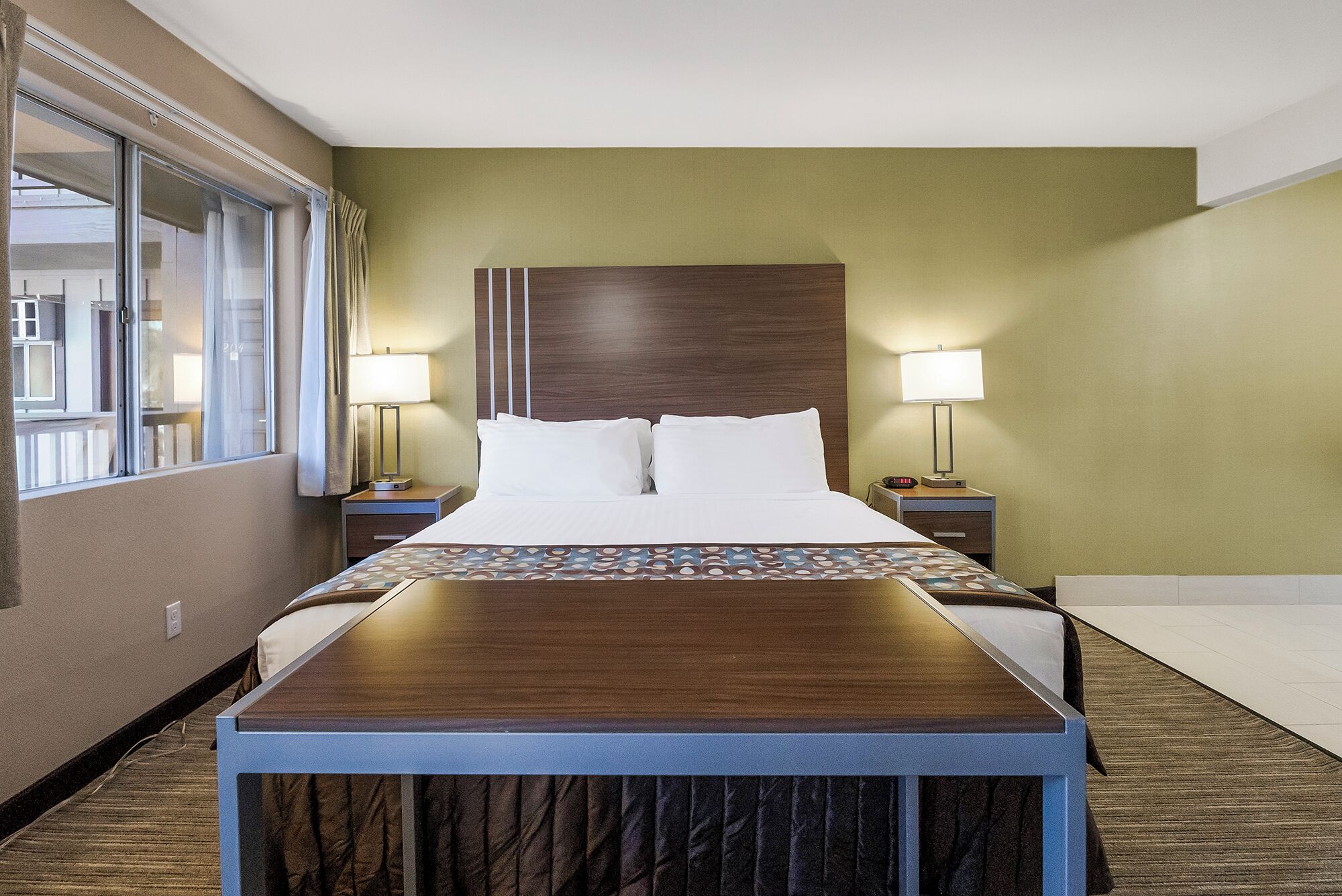 Een hotelkamer met een groot bed, twee nachtkastjes met lampen, een houten hoofdeinde en uitzicht naar buiten door een raam.