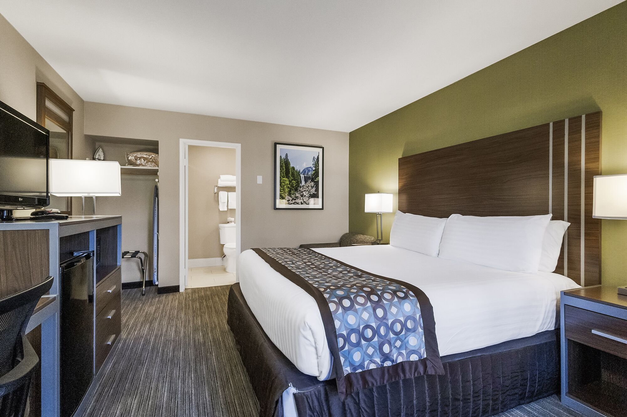 A hotel room with a bed, bedside tables, a desk with a TV, a mirror, and a bathroom visible through an open door.