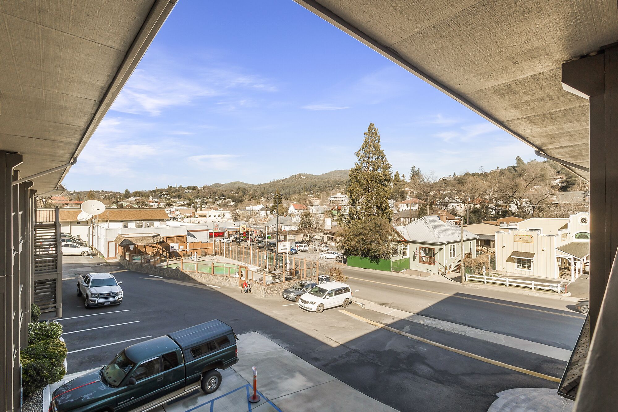 A view from under a bridge, overlooking a town with buildings, a large tree, and parked vehicles.