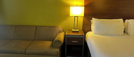 A hotel room with a sofa, a bedside table with a lamp, and a bed with pillows.
