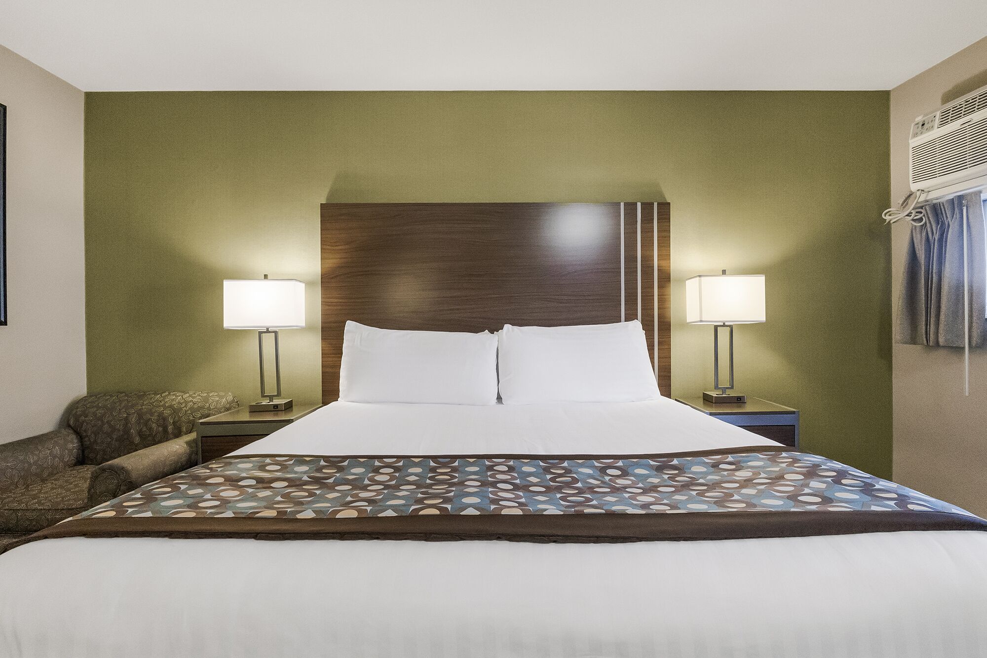 Een hotelkamer met een groot bed, twee bedlampjes, een bedspread met een patroon en een houten hoofdbord.