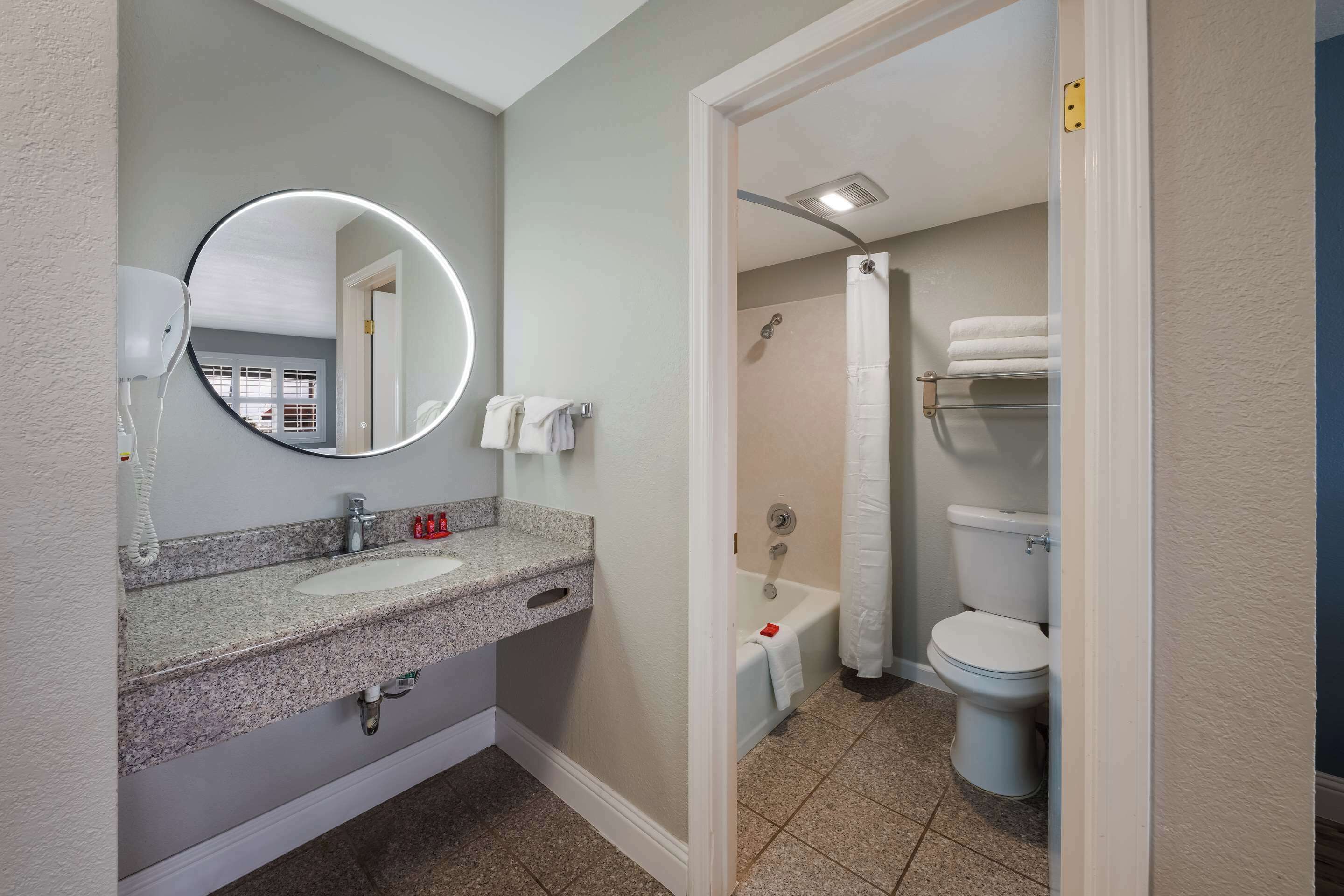 Un baño con lavamanos de granito, un espejo redondo con luz, ducha y retrete.