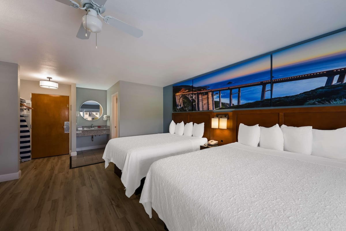 A hotel room with two beds, a ceiling fan, a painting of a bridge, and a wooden floor.