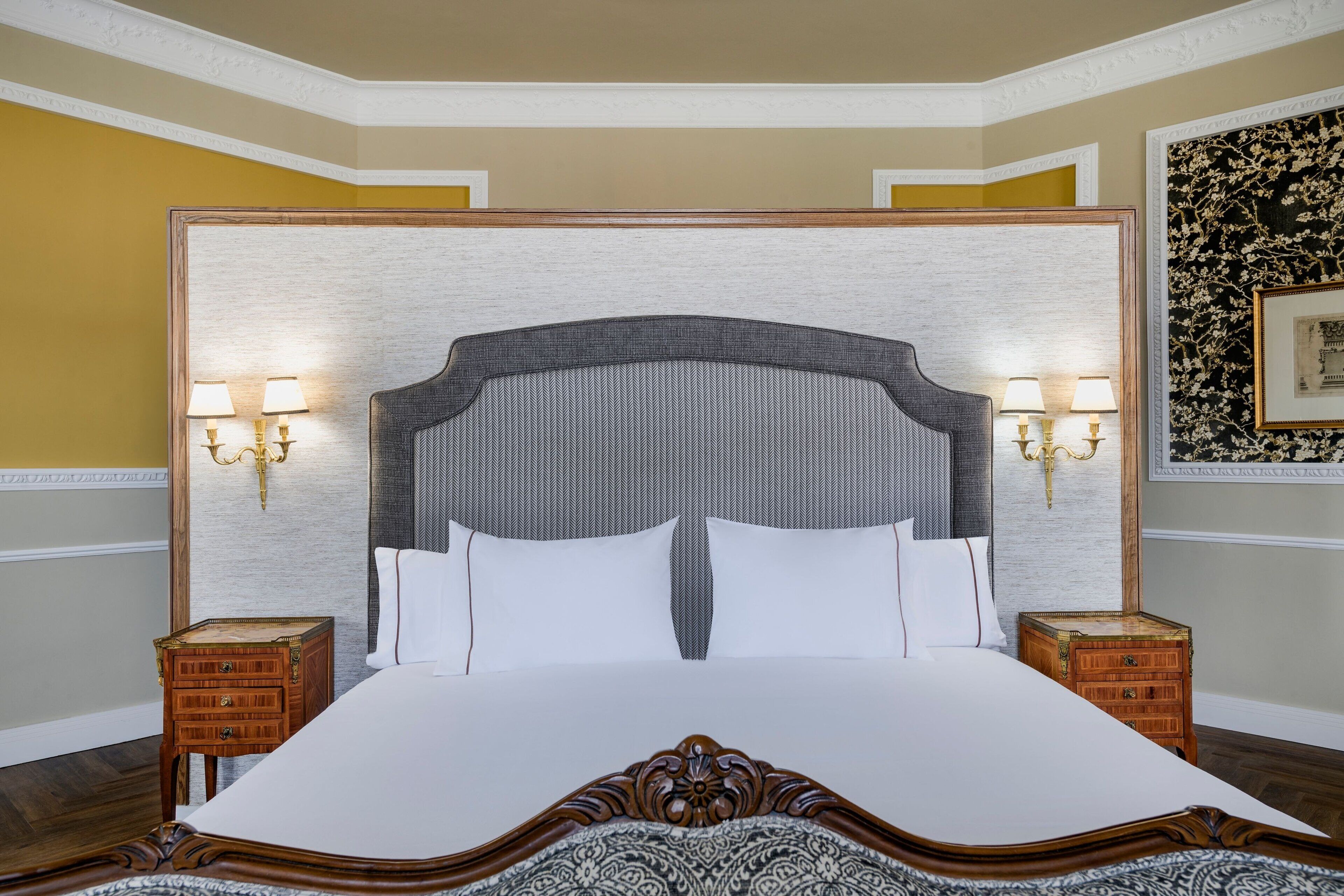 A large bed with a wooden headboard, two bedside tables with lamps, and a floral-patterned wall hanging.