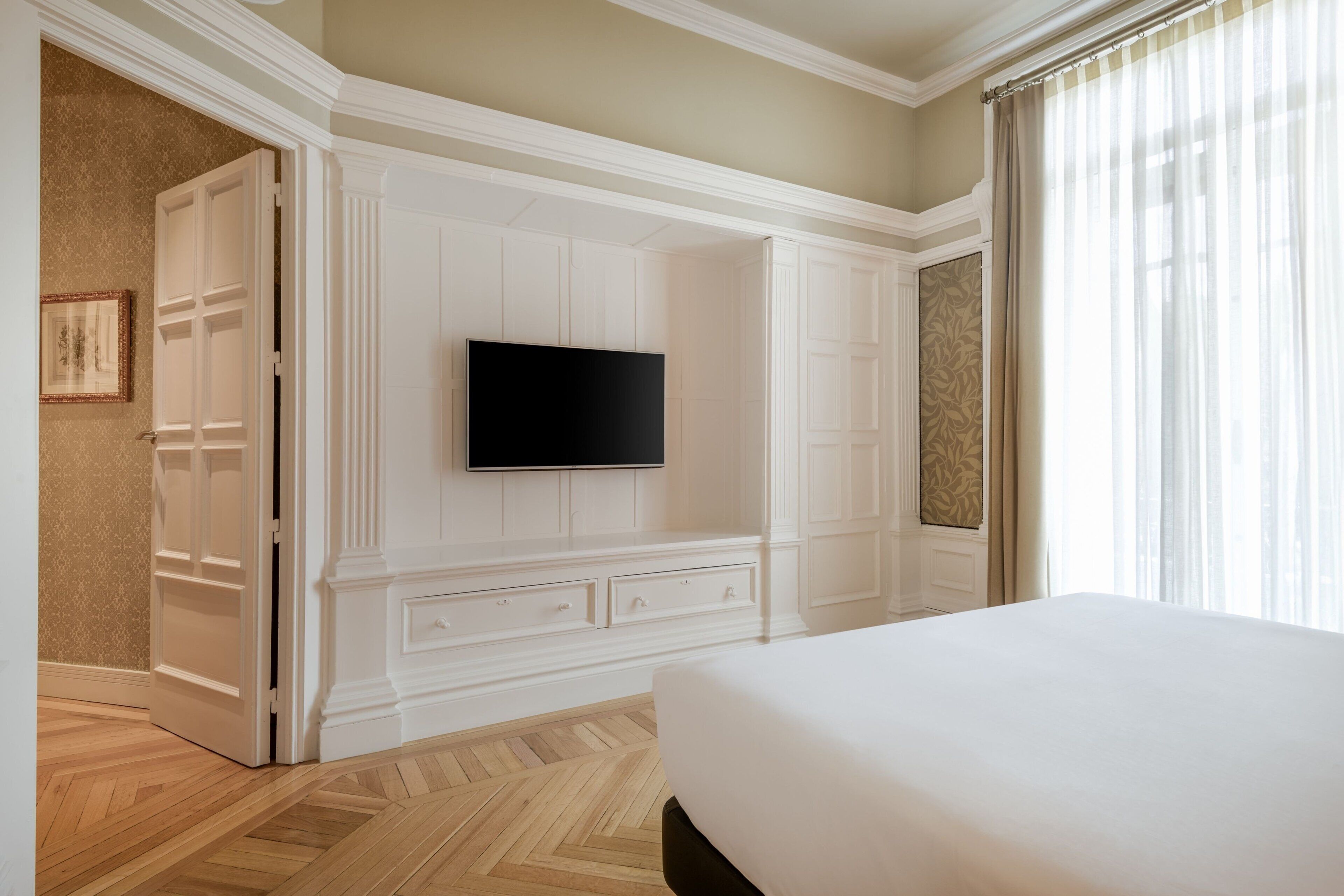 A hotel room with a large bed, a flat-screen TV mounted on the wall, a wardrobe with multiple drawers, and a door leading to another room.