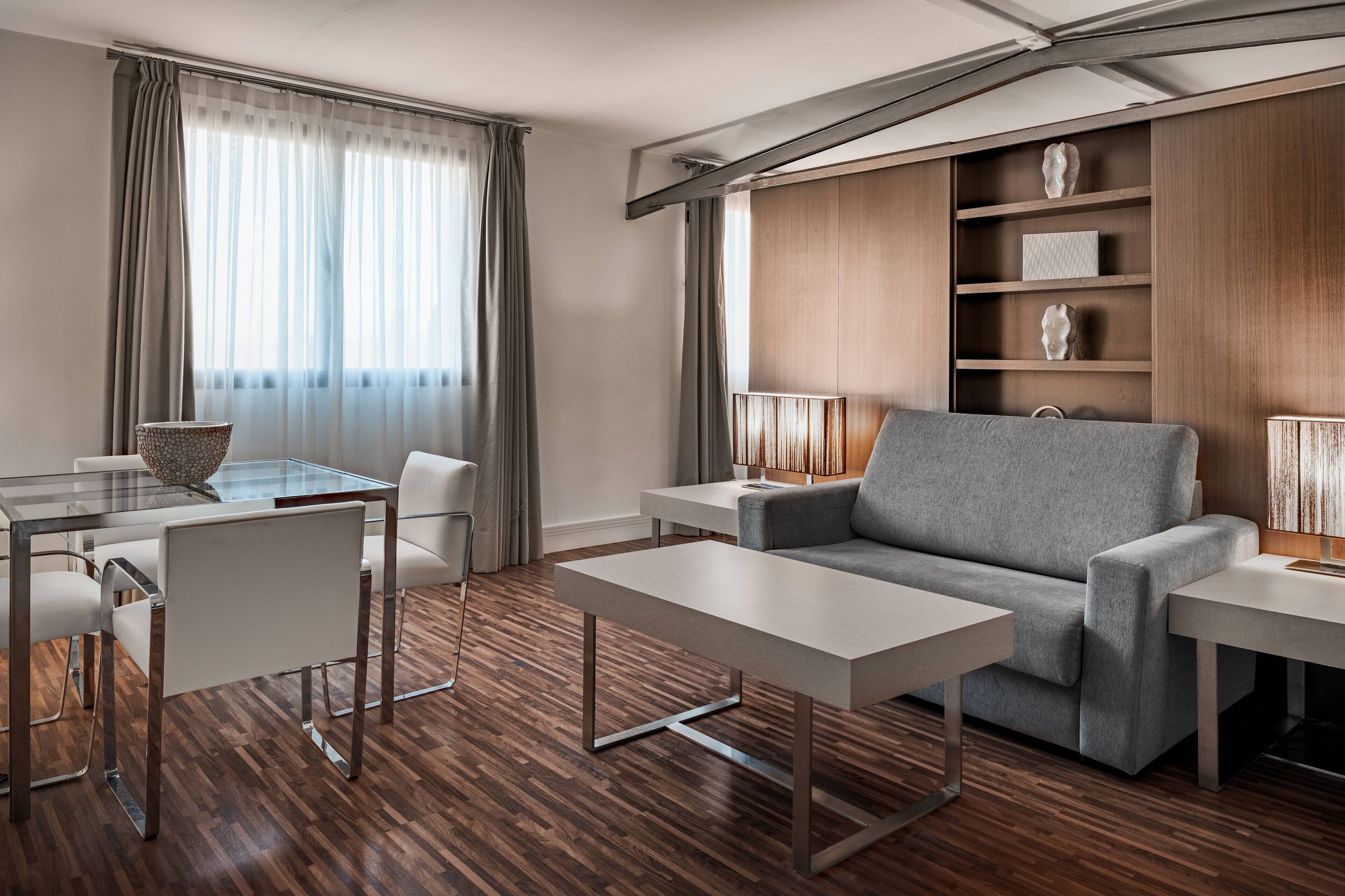 A modern living room with a glass dining table, white chairs, a grey sofa, and a wooden bookshelf.