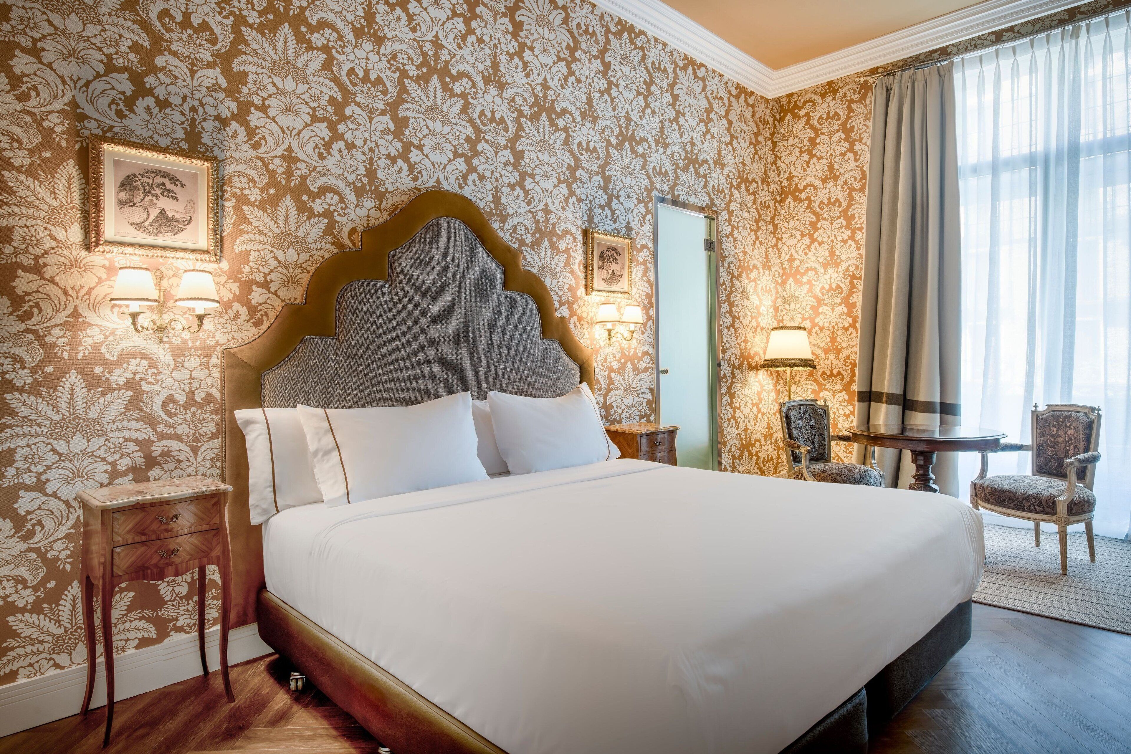 Una habitación de hotel con una cama grande, dos mesitas de noche, una butaca, una pequeña mesa y un espejo.