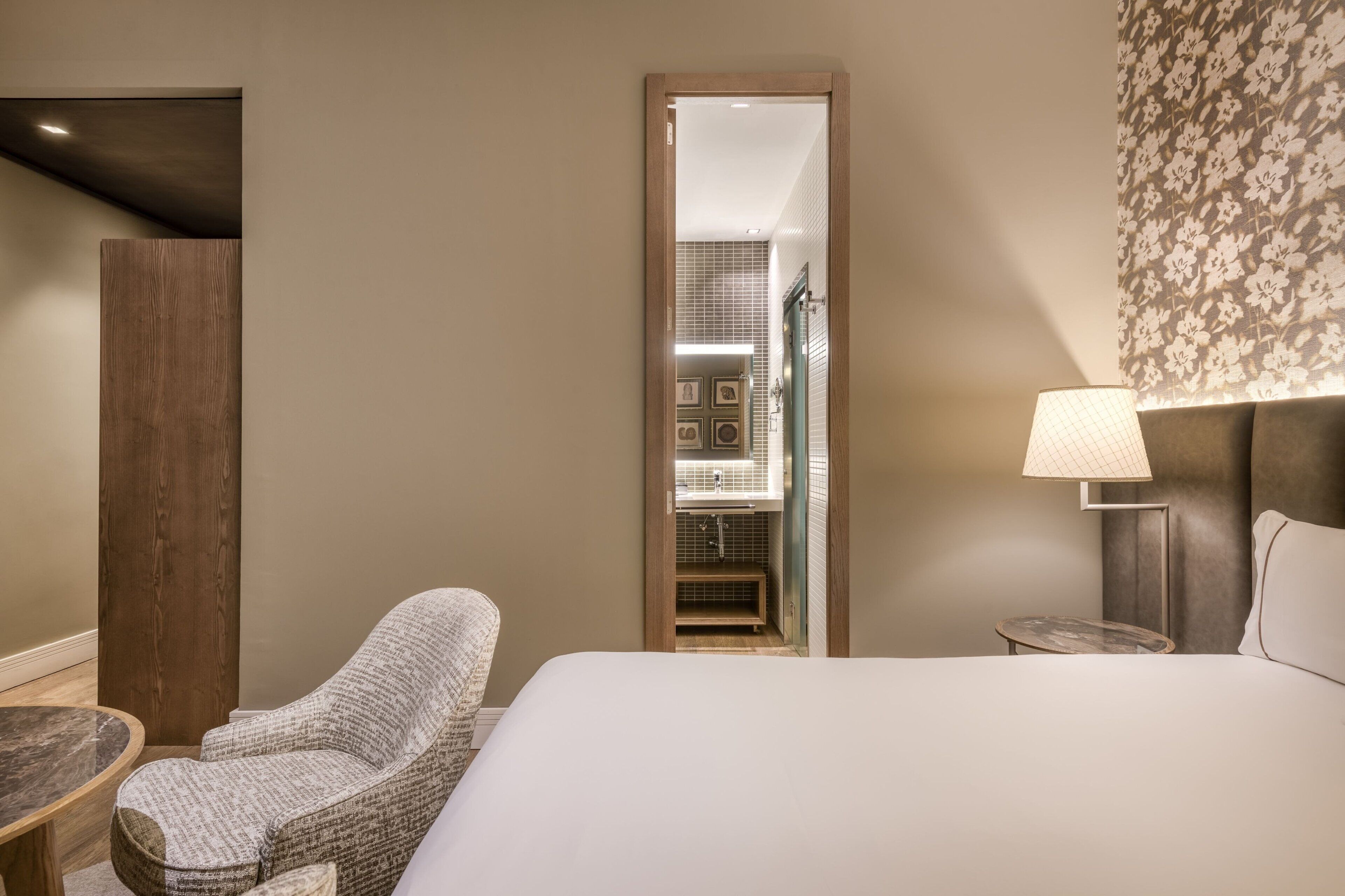 A hotel room with a bed, a chair, a small table, a lamp, and a mirror.