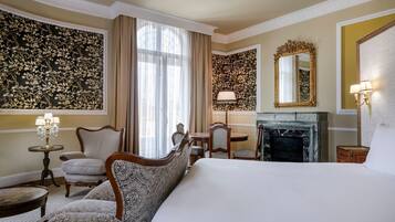 A hotel room with a large bed, a fireplace, a dining table with chairs, a chandelier, and a large mirror.