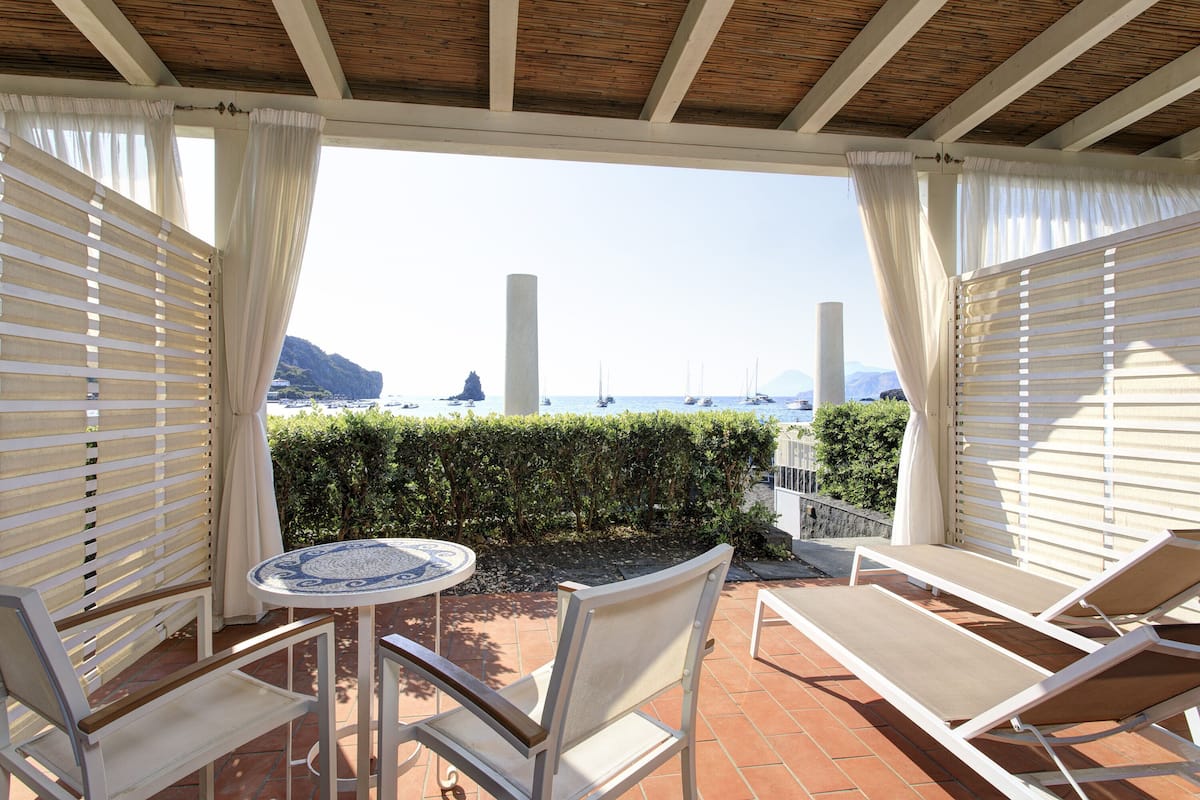 A covered patio with a table and two chairs, overlooking a coastal view with boats and a mountain.
