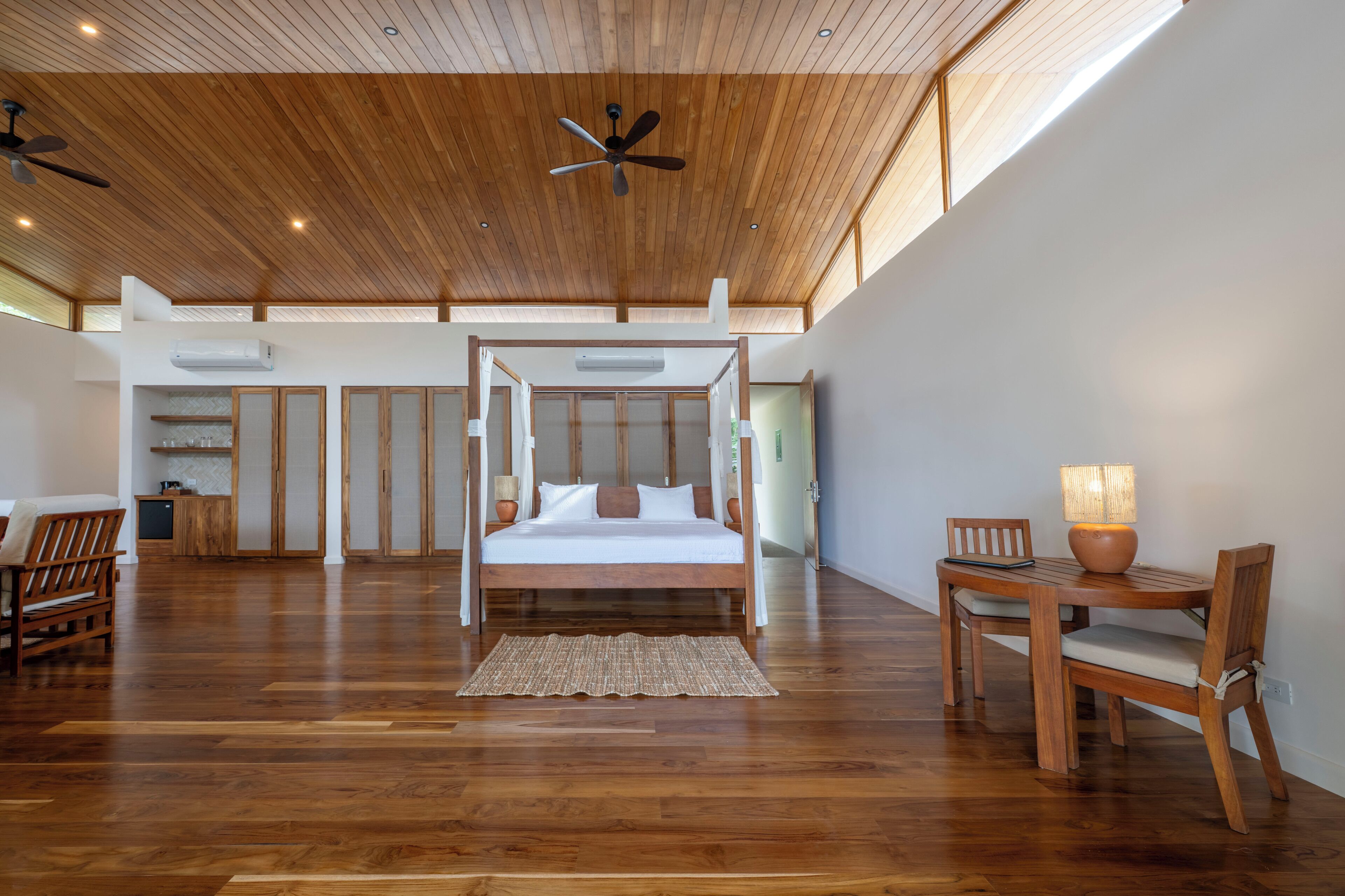 Un dormitorio amplio con techo de madera, una cama con dosel, una mesa de comedor con sillas y un ventilador de techo.