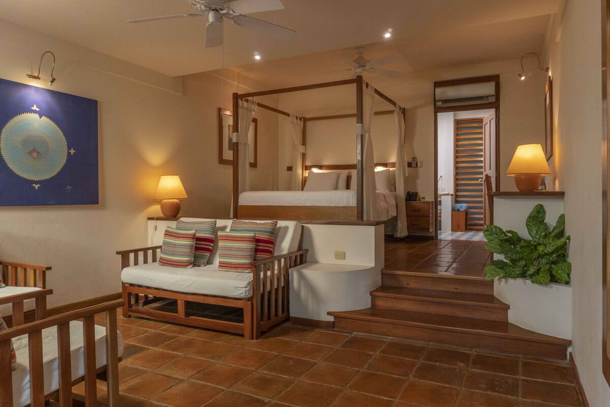A bedroom with a four-poster bed, a sitting area with a sofa and cushions, and a staircase leading to another room.