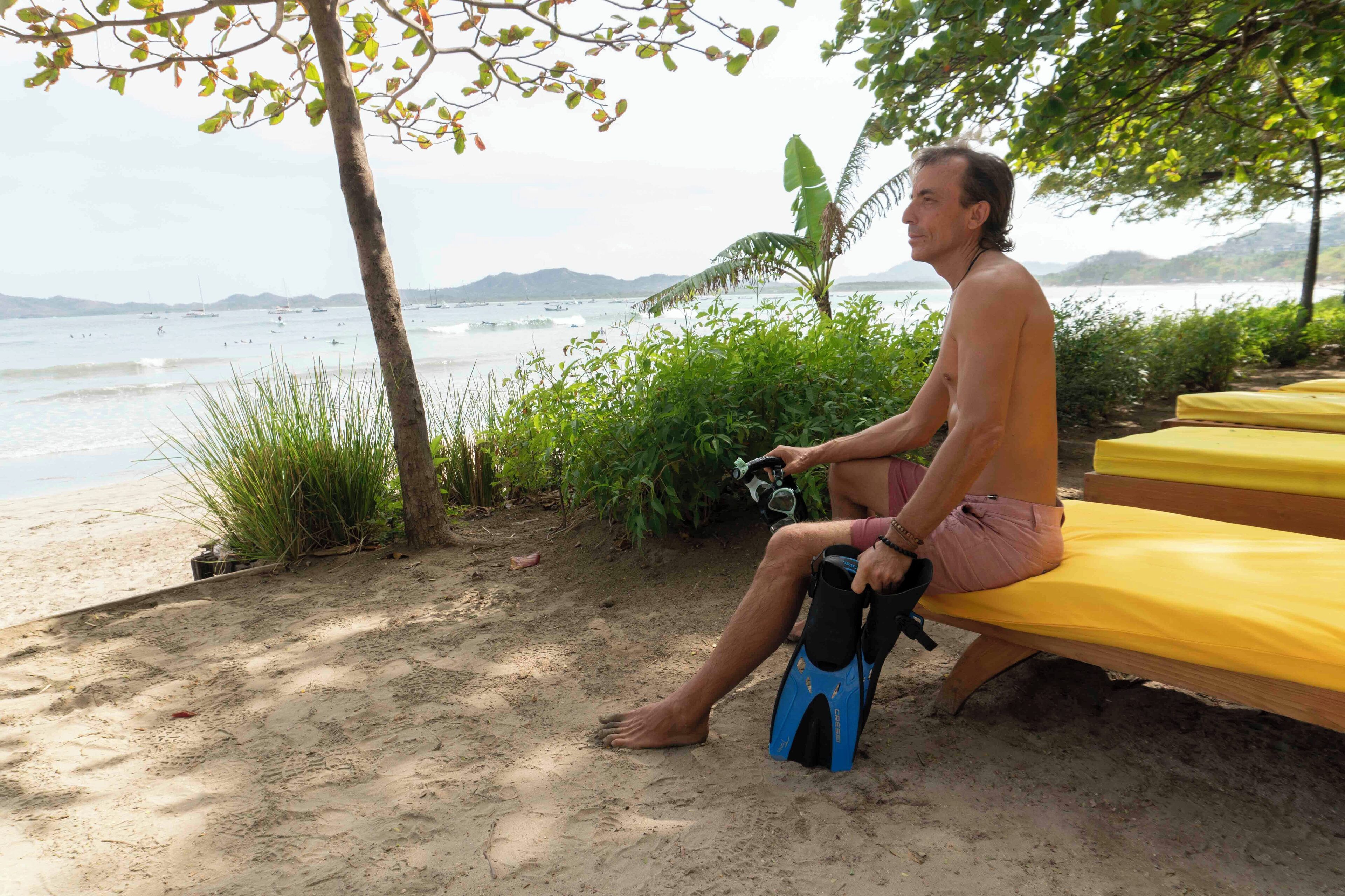 A person sitting on a yellow bench by the beach, holding a camera.