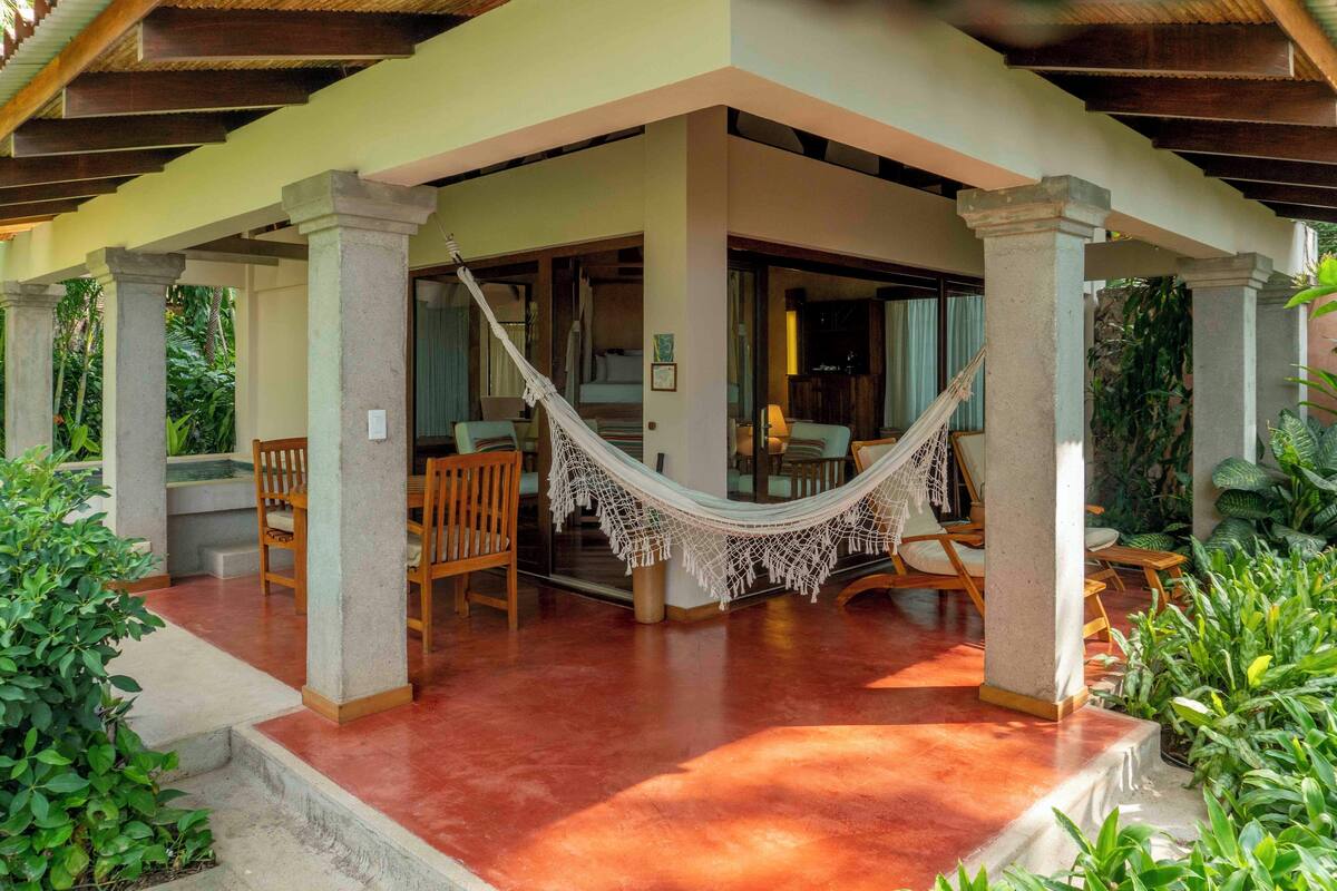 A covered outdoor seating area with hammocks, wooden chairs, and a concrete floor.