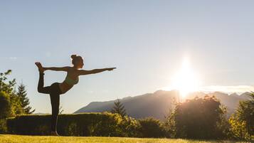 Una persona realizando una postura de yoga en un campo cubierto de hierba, con montañas al fondo.