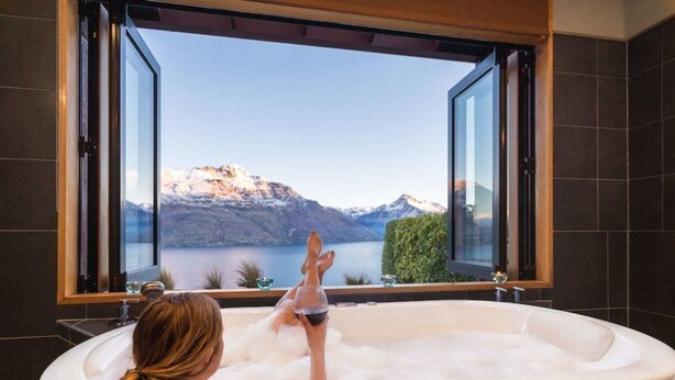 Una mujer se relaja en una bañera con vista a montañas y un lago a través de una ventana abierta.