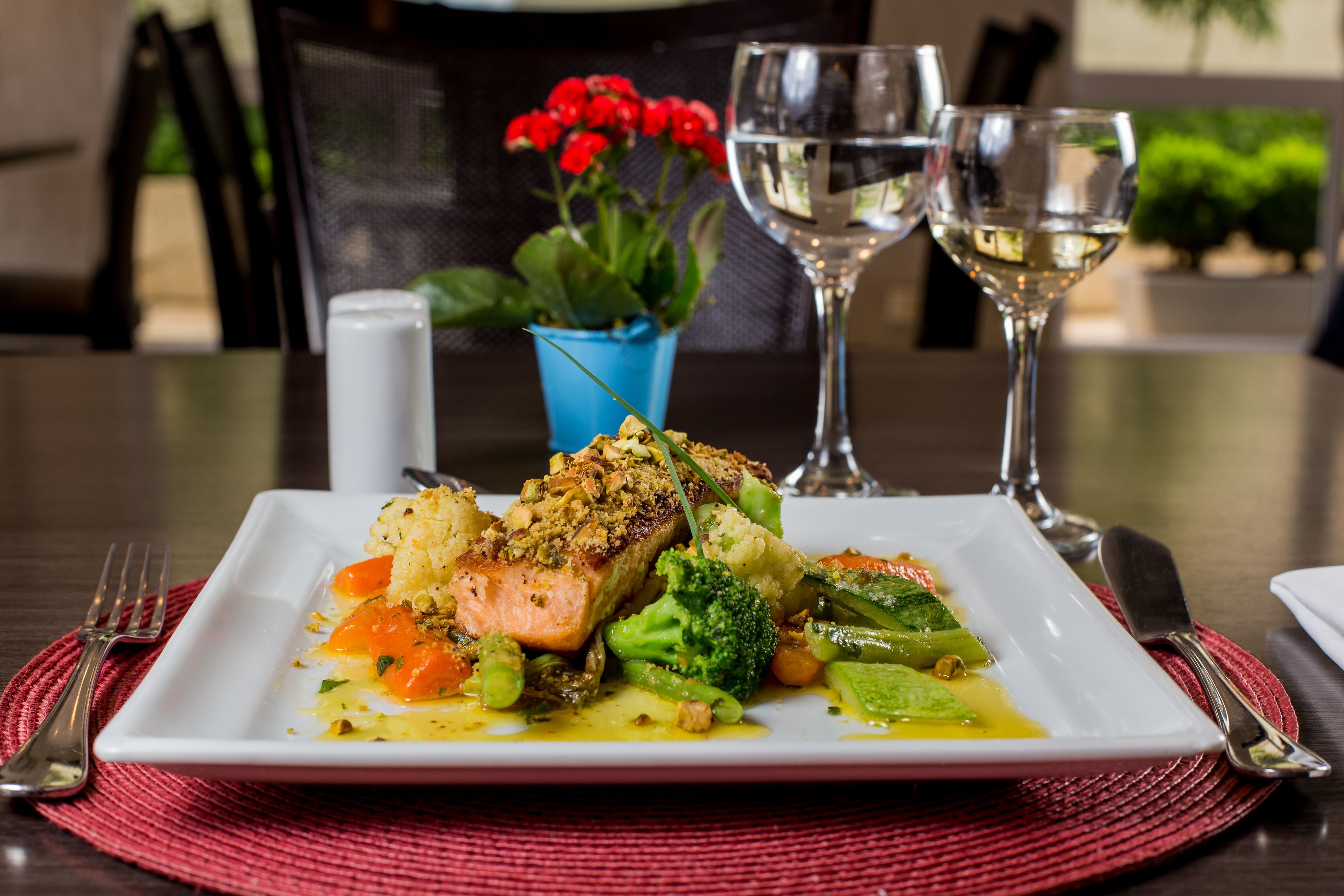 A square white plate with a piece of salmon, broccoli, carrots, and other vegetables, accompanied by two wine glasses and a blue flower vase on a table.