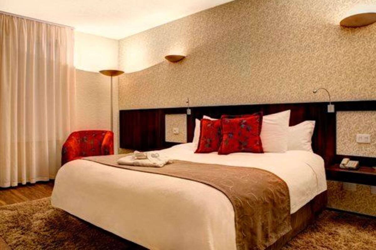 A hotel room with a large bed, a red armchair, a nightstand, and a wall-mounted lamp.