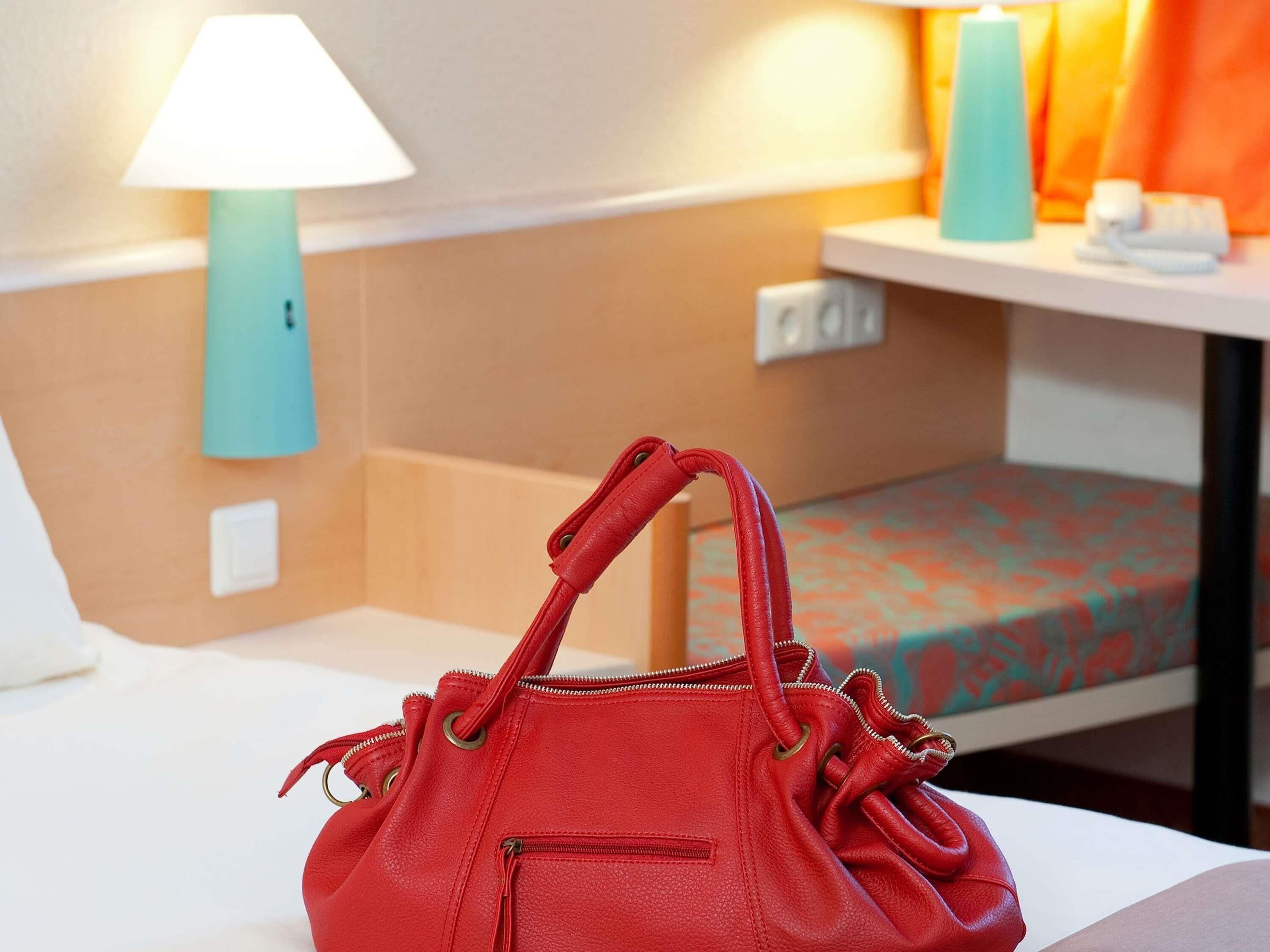 A red handbag on a bed with a patterned bedspread, a bedside table with a lamp, and a wall socket.