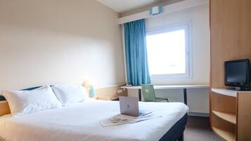 A hotel room with a bed, a desk with a laptop, a TV, and a window with curtains.