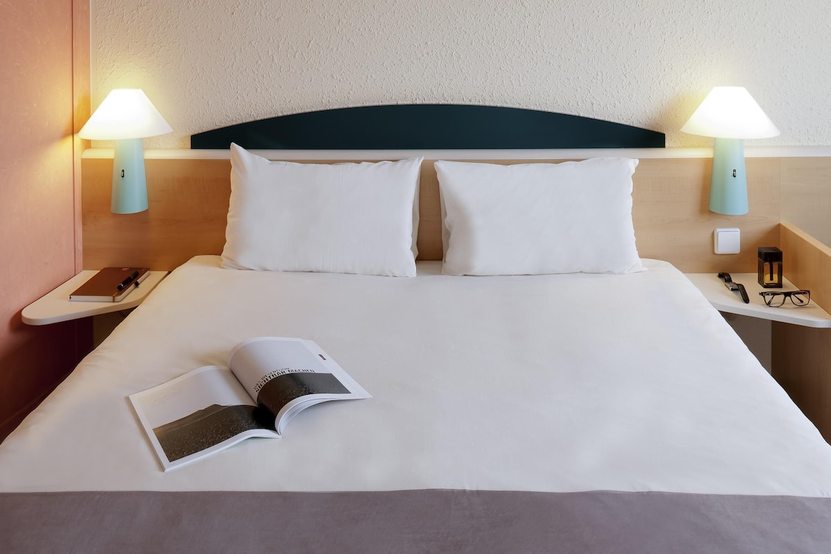 A neatly made bed with white linens, two bedside lamps, a nightstand with a phone, glasses, and a book.