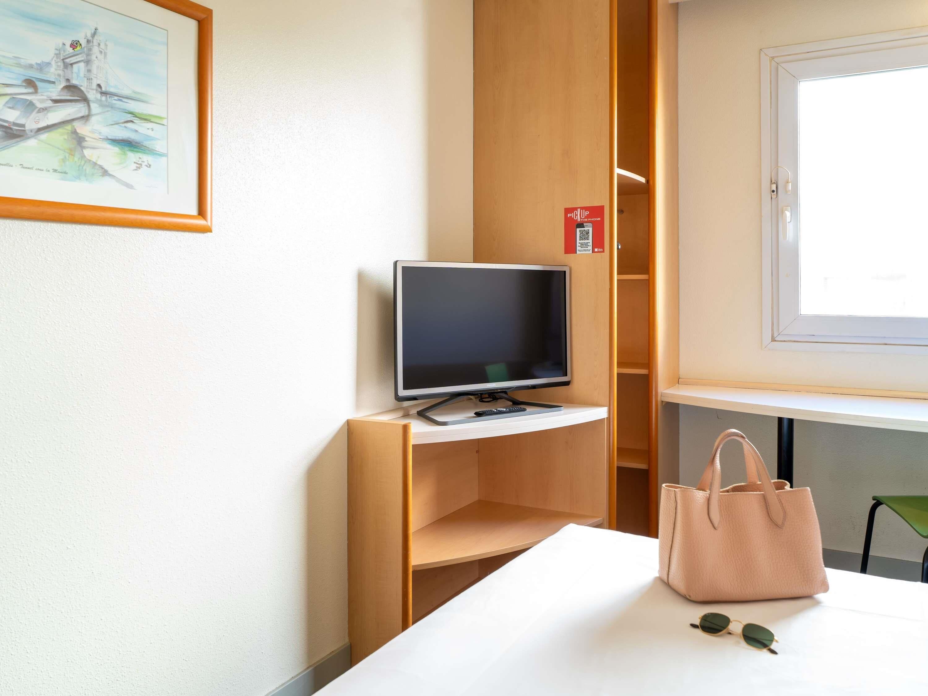 A hotel room with a television on a wooden stand, a painting of a bridge, a window, and a table with a handbag and sunglasses.