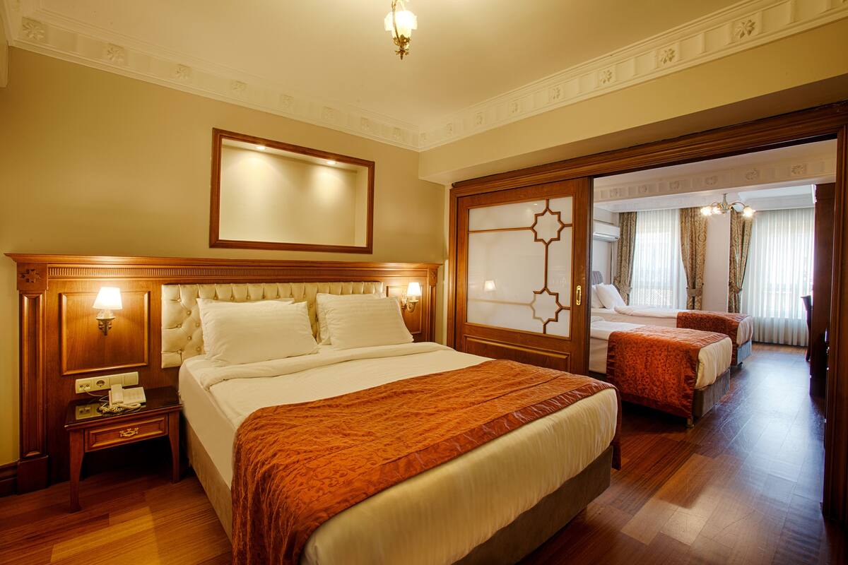 Habitación de hotel con una cama grande, dos mesitas de noche con lámparas, un espejo sobre la cama y una puerta corredera que da a otra habitación.